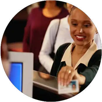 A young Black woman in business attire smiles and holds up her driver’s license to a DMV worker.