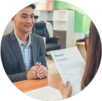 A young Asian man sits at a desk and smiles as a woman sits across from him and looks at his resume.