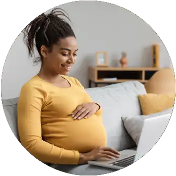 A young pregnant Black woman smiles and works on a laptop while sitting on a couch in a living room.