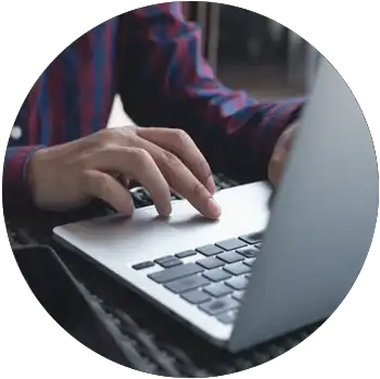 The hands of a man with tan skin and a checkered button-down shirt work on a laptop.